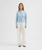 Woman wearing a light blue ribbed cardigan over a white top with white pants on a light gray background